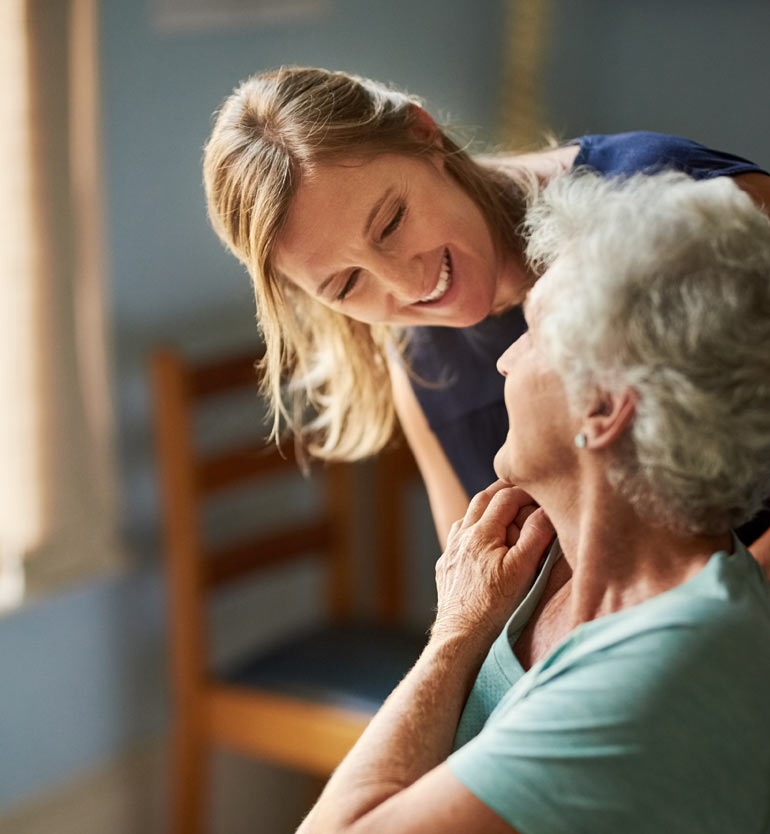 _-senior-memory-care-caregiver- Caregiver smiling and interacting with a senior woman in a memory care unit.