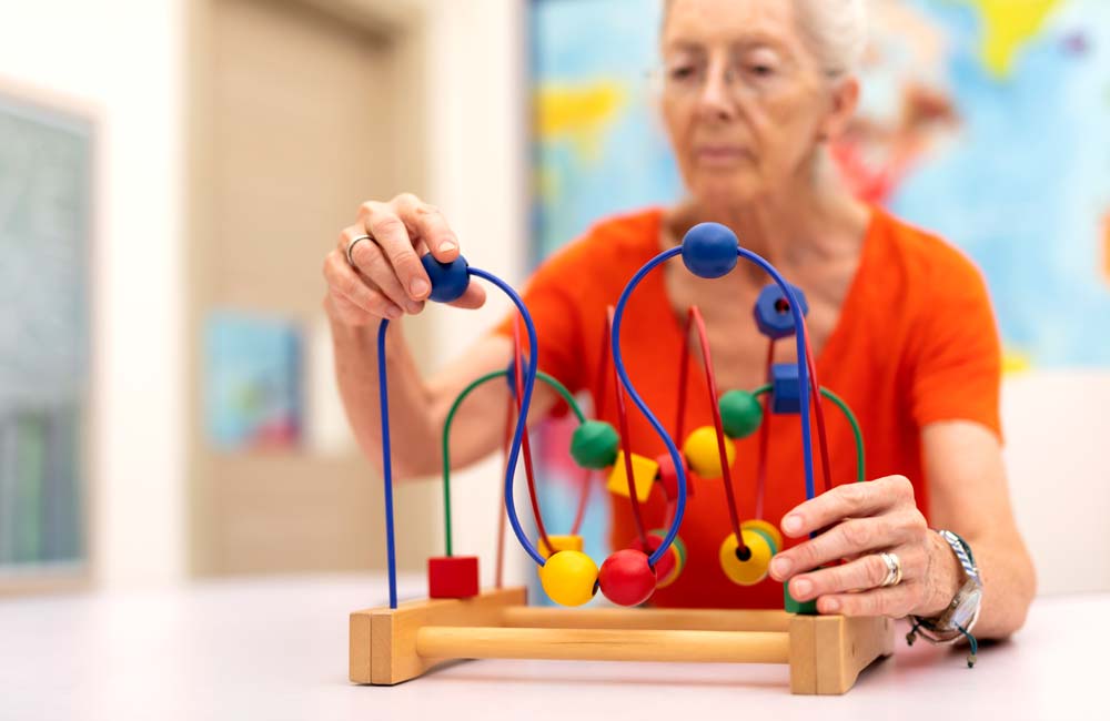 senior-memory-care--activity Senior woman engaging in a memory care activity with a colorful bead maze at a senior living unit.