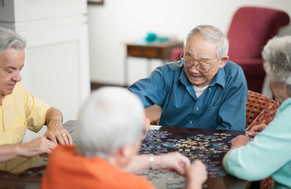 seniors-doing-puzzle Elderly residents working together on a puzzle at a shared table in a living space.