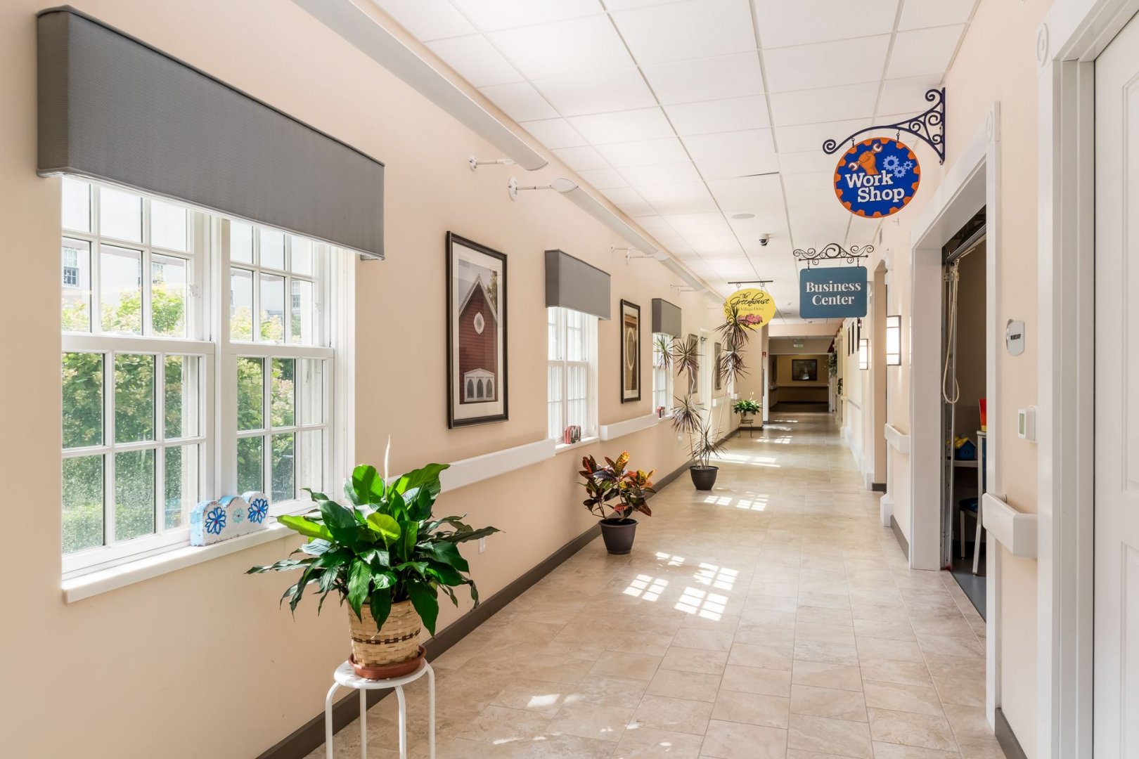 Bright Hallway in Senior Living Community Sunny hallway with plants, large windows, and signs for a business center and workshop.